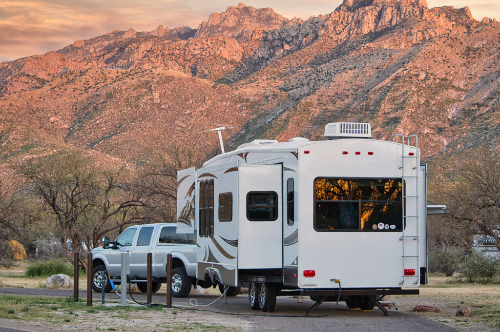 A pickup truck is parked with a large white camper trailer at a campsite, with rugged mountains in the background at sunset.