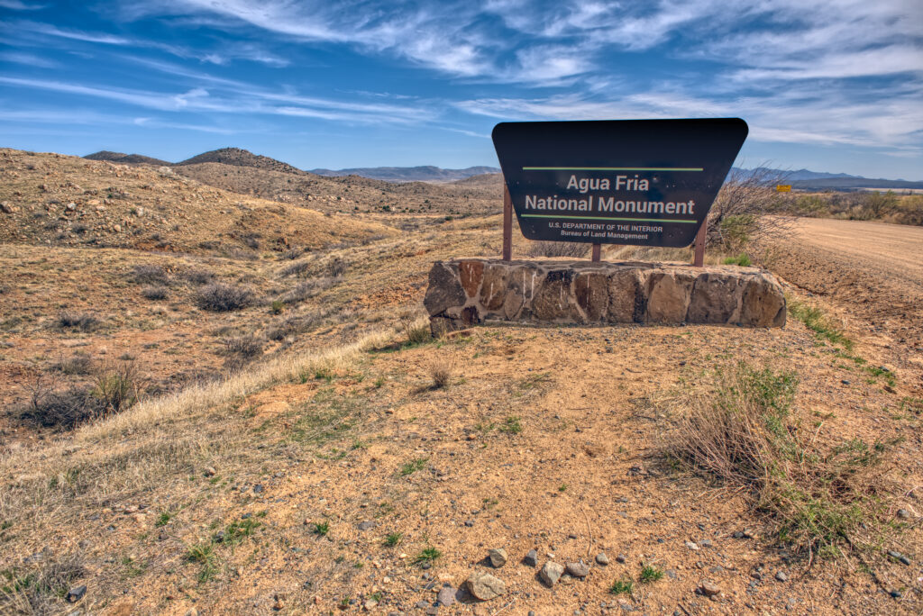 A stone sign reads "Agua Fria National Monument" beside a dirt road in a dry, rocky desert landscape under a blue sky.