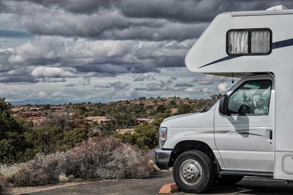 A white RV is parked near a desert landscape under a cloudy sky, with a dog visible in the passenger seat window.