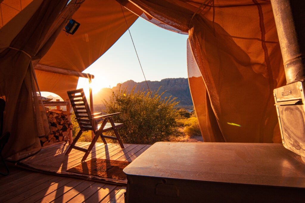 View from inside a canvas tent showing a wooden chair, firewood, stove, and sunrise over a landscape with mountains and bushes.