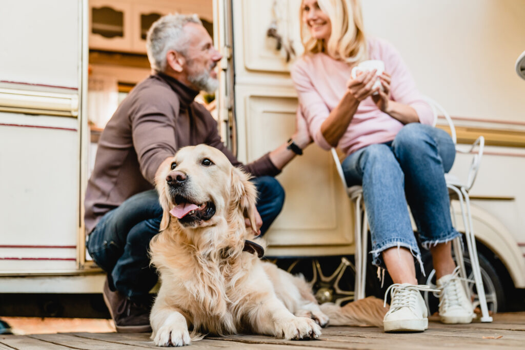 A golden retriever lies on a wooden deck in front of a camper while a man crouches beside it and a woman sits on a chair, both smiling and talking.