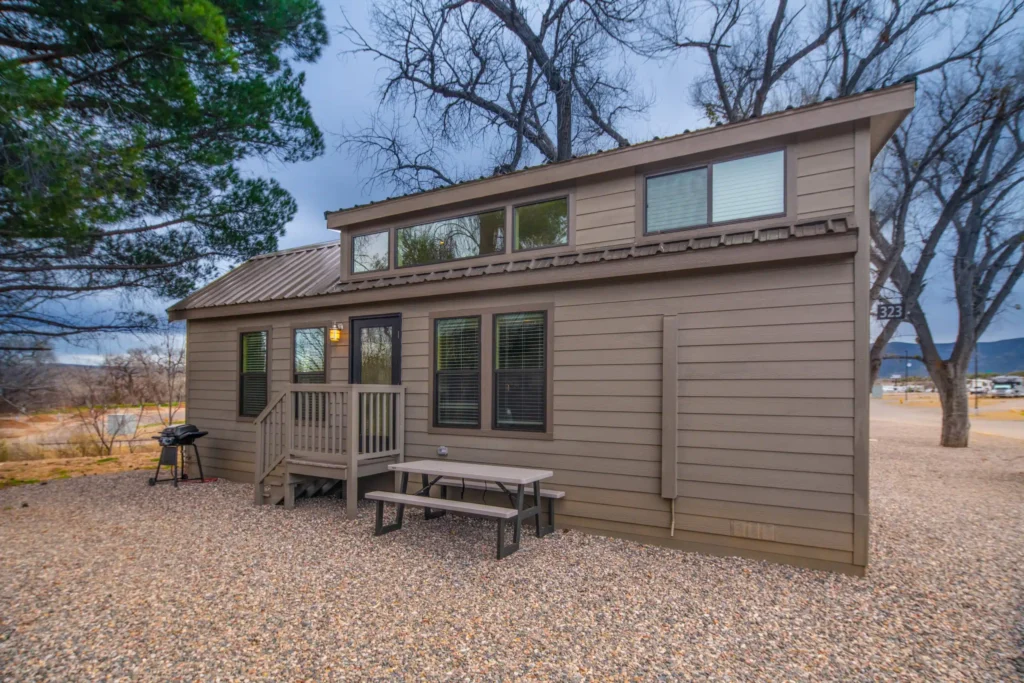 A small, modern tiny house with a raised porch, windows along the top, picnic table, and grill, set on a gravel lot with trees and cloudy sky in the background.