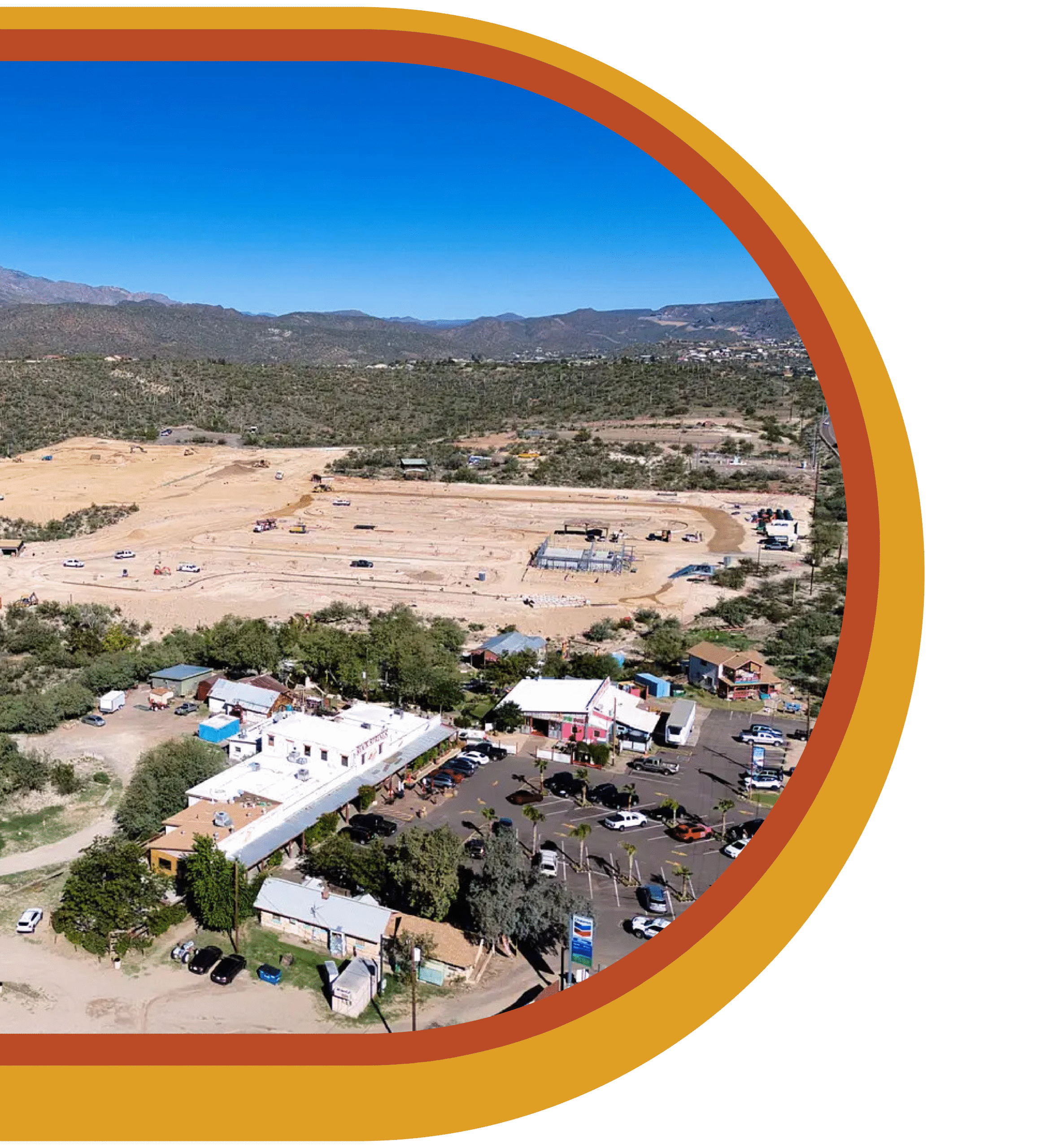 Aerial view of a small town with scattered buildings, parked cars, and construction areas, surrounded by desert landscape and mountains under a clear blue sky.