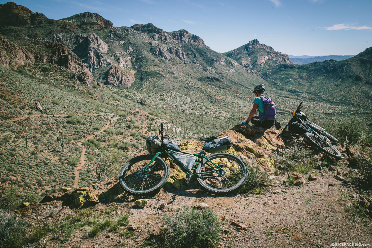 A person sits on a rocky ledge next to two bicycles, overlooking a vast, rugged mountain landscape under a clear sky.