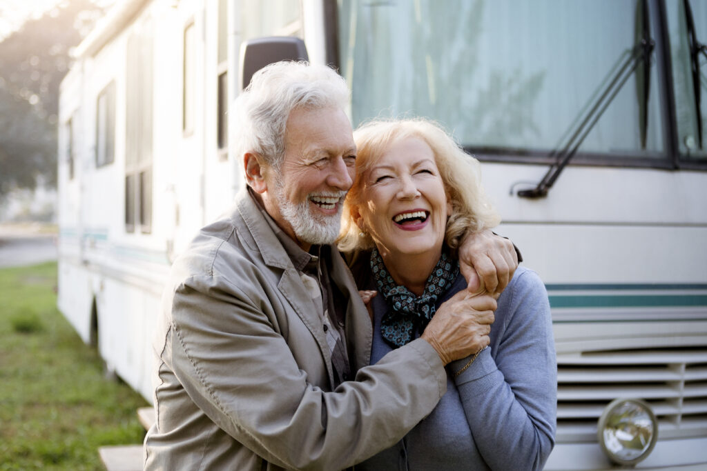 Smiling older couple standing outside and embracing in front of a white RV during daytime.