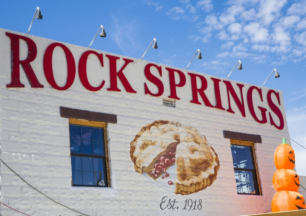 The side of a white building reads “ROCK SPRINGS” with a painted pie, two windows, “Est. 1918,” and stacked inflatable pumpkins, under a blue sky with clouds.