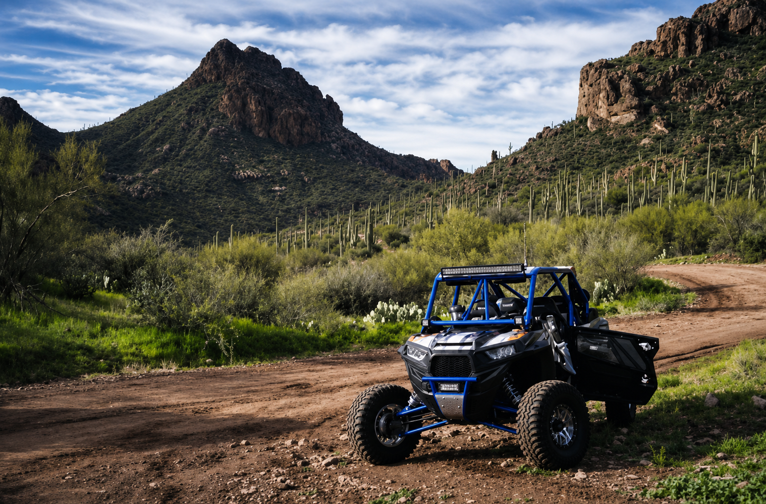 A blue off-road vehicle parked on a dirt path in a desert landscape with mountains, green vegetation, and scattered saguaro cacti under a partly cloudy sky.