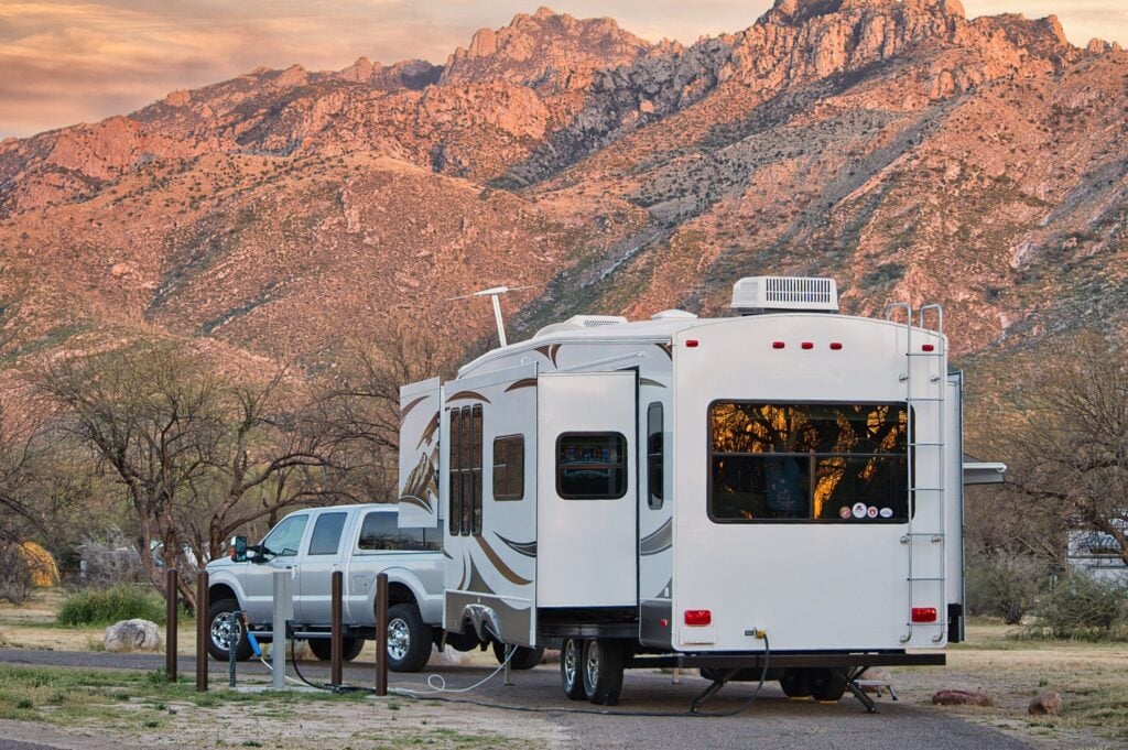 A silver pickup truck towing a white RV is parked at a campsite with mountains and trees in the background during sunset.
