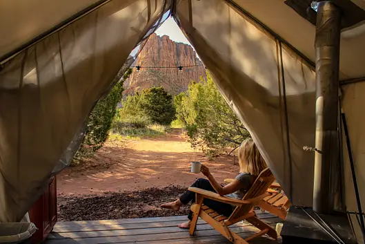 A person sits in a wooden chair inside a tent, holding a mug and looking out at trees and a rocky mountain in the distance.