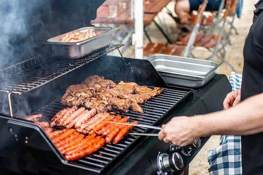 Person grilling sausages and marinated meat on a barbecue grill outdoors, with a tray of shrimp on the upper rack and picnic tables in the background.