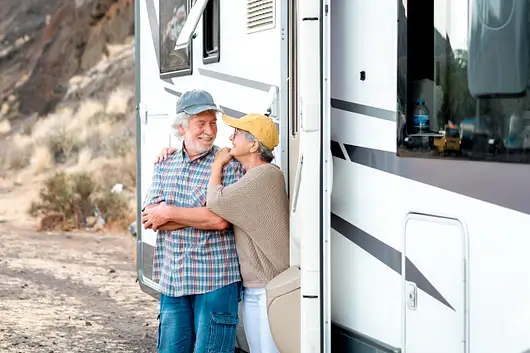 An older couple stands next to a parked RV in a rocky outdoor setting, smiling at each other.