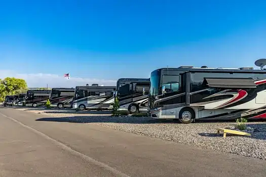 Several large RVs are parked side by side on gravel lots under a clear blue sky, with an American flag visible in the background.